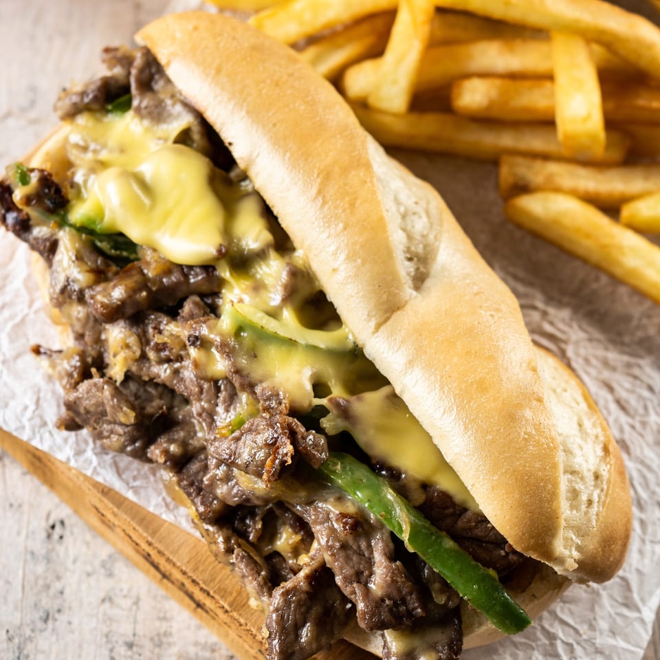 A Corner Cafe cheesesteak sandwich with melted cheese, green peppers, and fries on parchment paper.